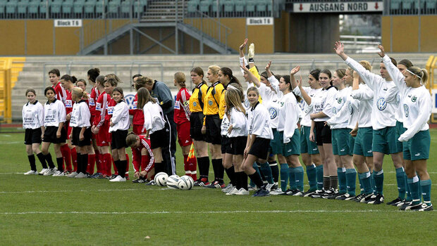 Die ehemalige Frauenmannschaft des VfL Wolfsburg steht mit der gegnerischen Mannschaft in einer Reihe und winkt.