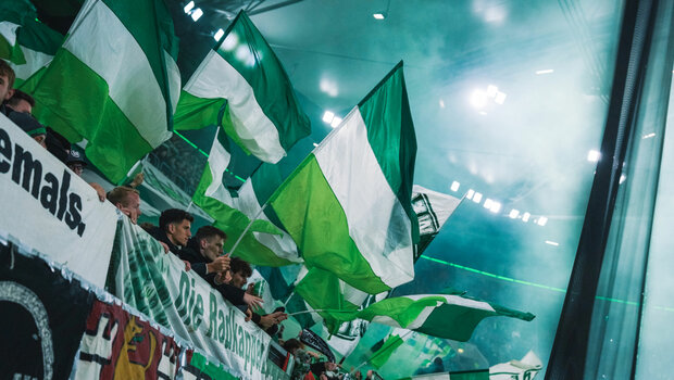 Fans des VfL Wolfsburg schwenken in der Nordkurve grün-weiße Fahnen.