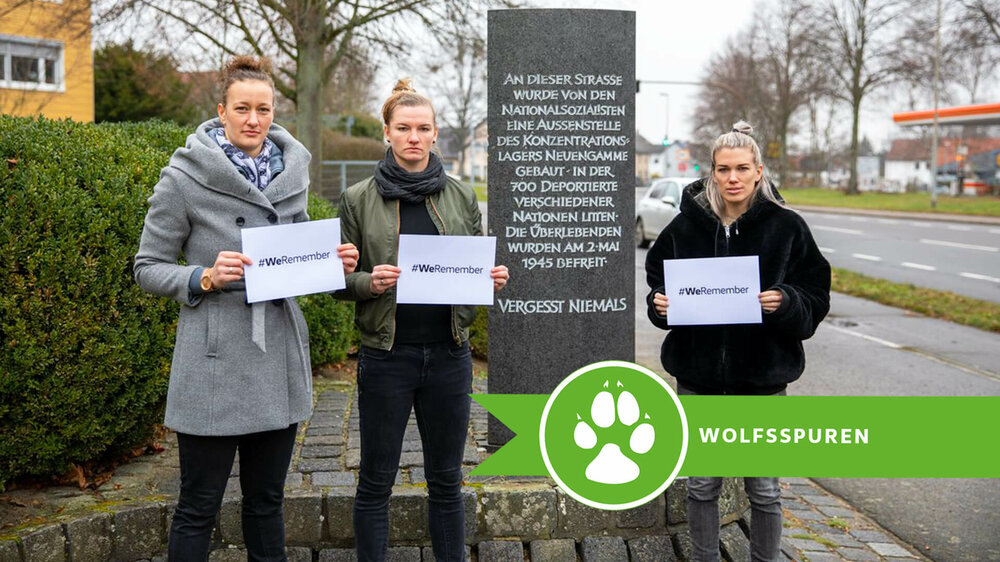 VfL Wolfsburg Spielerinnen posieren mit #WeRemember Schildern vor einem Denkmal.