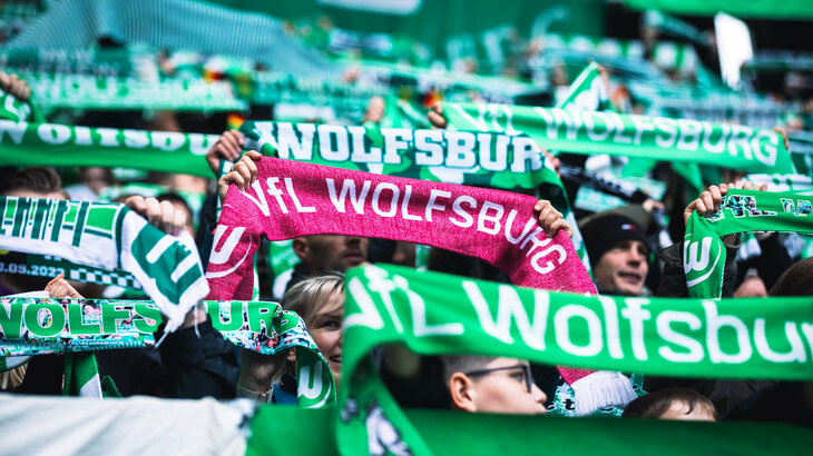 Die Fans des VfL Wolfsburg halten ausgebreitete Fanschals nach oben.