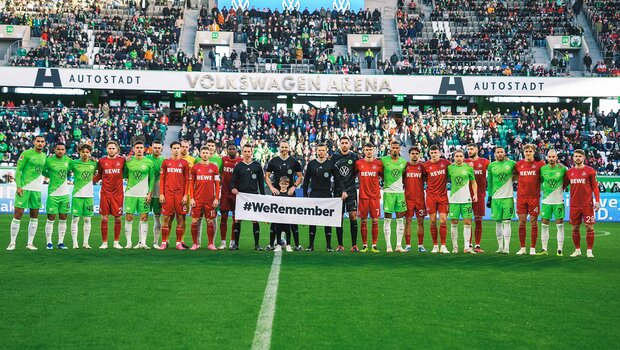 Die Mannschaften des VfL Wolfsburg und des 1. FC Köln gemeinsam mit den Schiedsrichtern und dem Banner #WeRemember.