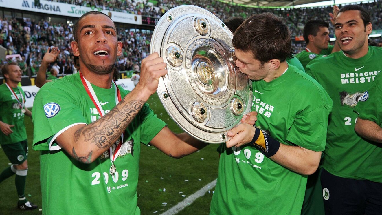Der ehemalige Spieler des VfL Wolfsburg Ashkan Dejagah steht auf dem Spielfeld und hält gemeinsam mit einem seiner Mitspieler die Meisterschale in der Hand.