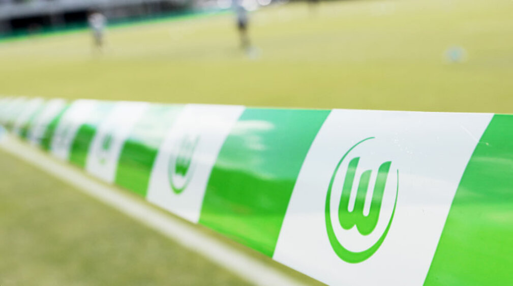 Ein VfL Wolfsburg-Flatterband ist im Vordergrund, im Hintergrund erkannt man unscharf einen Fußballplatz.
