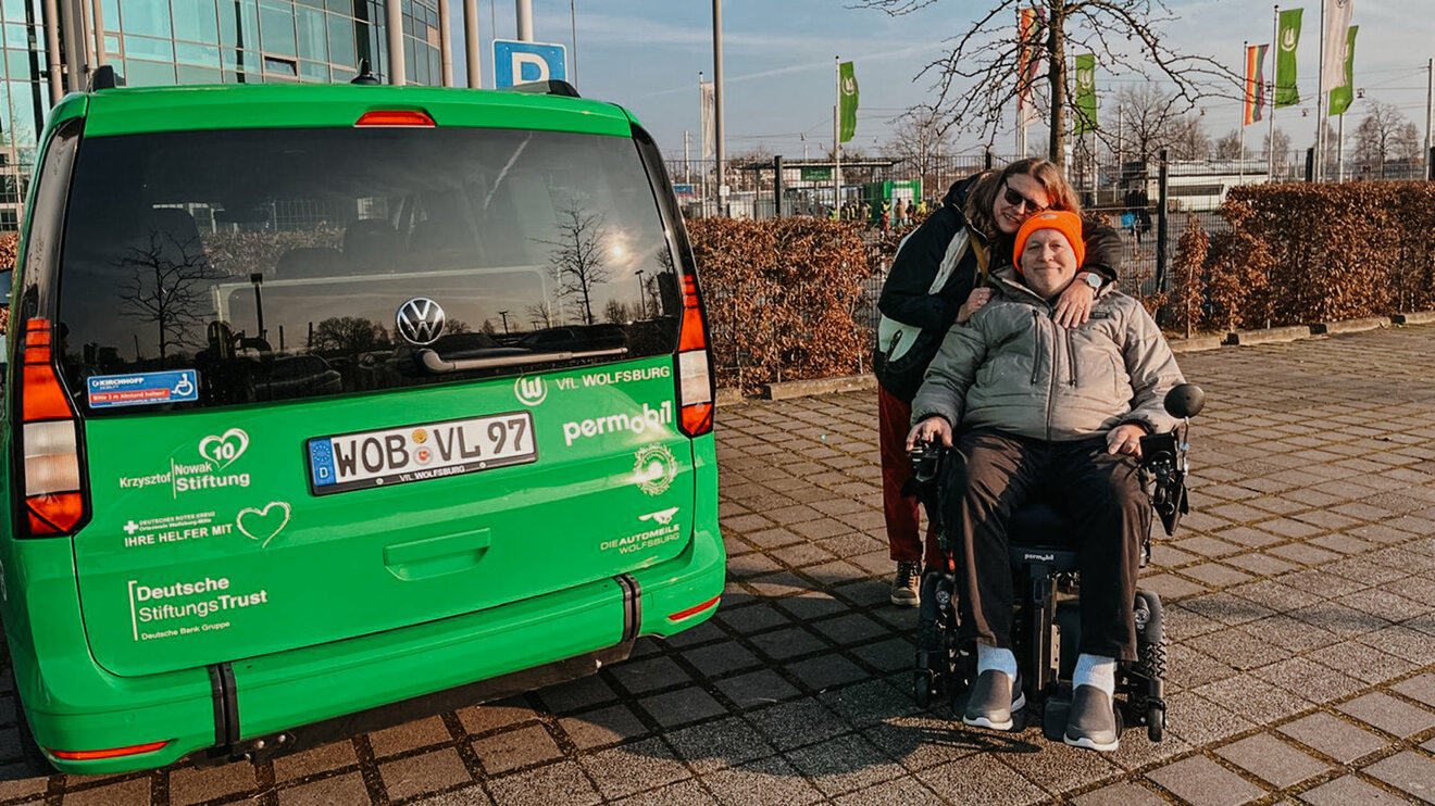 Eine ALS erkrankte Person befindet sich mit Rollstuhl vor der Volkswagen Arena und lächelt für ein Foto in die Kamera.