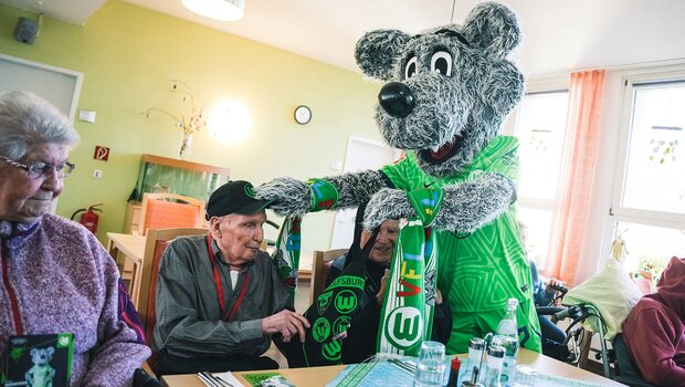 VfL-Wolfsburg-Maskottchen Wölfi übergibt einem AWO-Altenheim-Bewohner eine Tasche und eine Mütze.