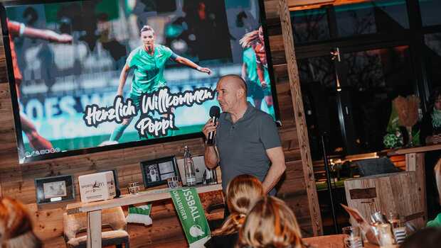 Sven Froberg hält in der rechten Hand das Mikrofon und spricht zu den Fans die zum Fantalk gekommen sind. Im Hintergrund ist eine Leinwand, auf der VfL Spielerin Alexandra Popp zu sehen ist und der Schriftzug Herzlich Wilkommen Poppi.
