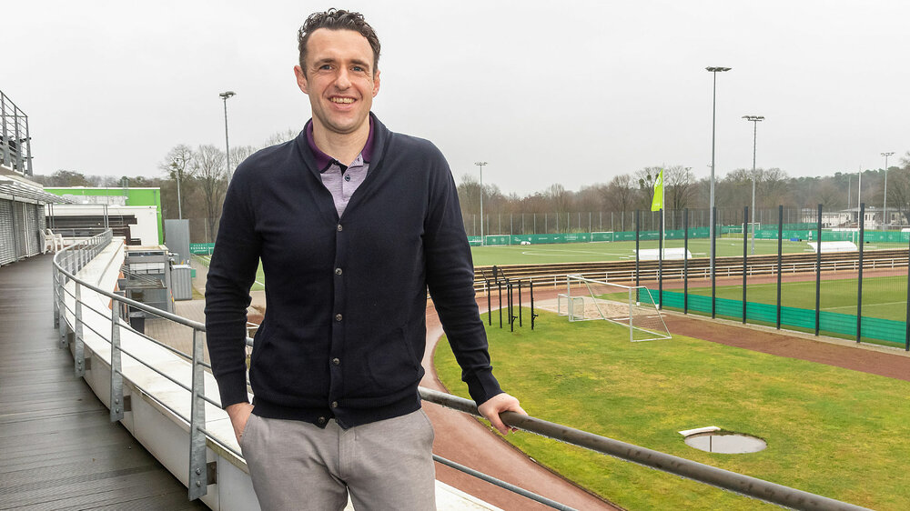Michael Gentner steht auf dem Balkon der VfL-Akademie. Im Hintergrund sieht man die Trainingsplätze der Nachwuchsmannschaften des VfL Wolfsburg.