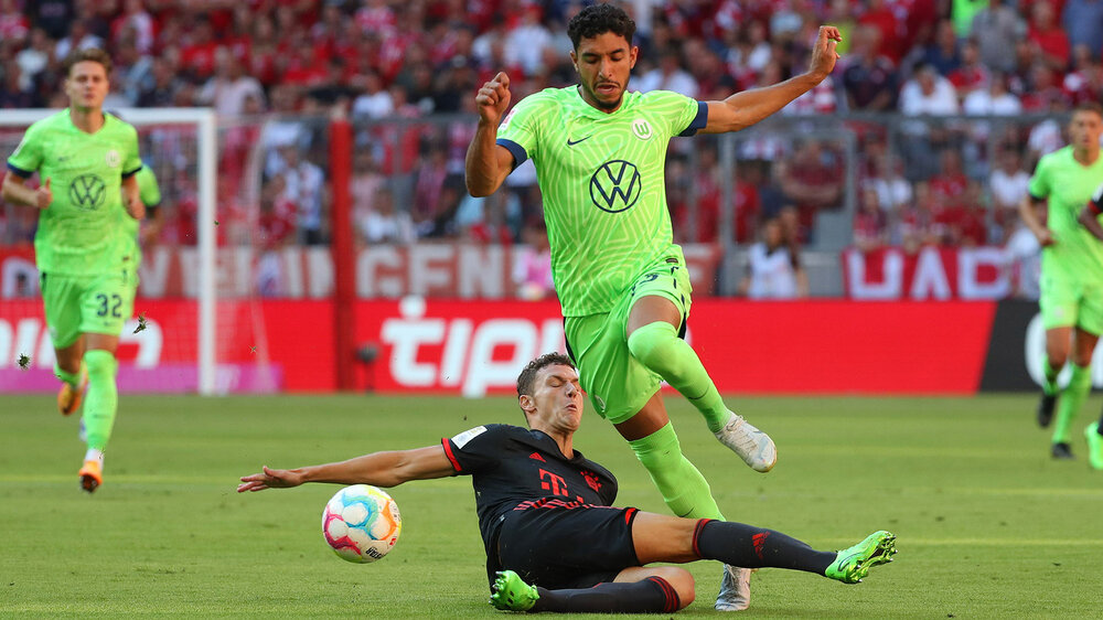VfL-Wolfsburg-Spieler Omar Marmoush springt über einen Gegenspieler herüber und läuft hinter dem Ball her.