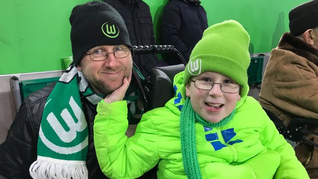 Ein junger VfL Wolfsburg-Fan und sein Vater lachen in die Kamera.