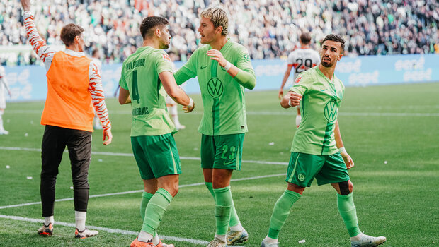 Die VfL-Wolfsburg-Spieler Koulierakis, Wind und Amoura stehen auf dem Rasen. Dabei kaltschen sich Koulierakis und Wind ab, während Amoura die Faus in die Lust streckt.