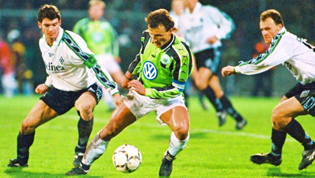 Spielsituation. Am Ball ein VfL-Wolfsburg-Spieler. Der Ball ist zwischen seinen beiden Füßen, der Spieler ist von vorne zu sehen. Von links und rechts kommend zwei Gladbach-Spieler, die versuchen den Ball zu bekommen.