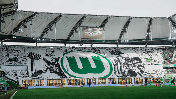Die Nordkurve in der Volkswagen Arena zeigt eine Choreografie. In der Mitte ist das VfL-Logo, rundherum sind viele ehemalige Spieler des VfL Wolfsburg zu sehen.