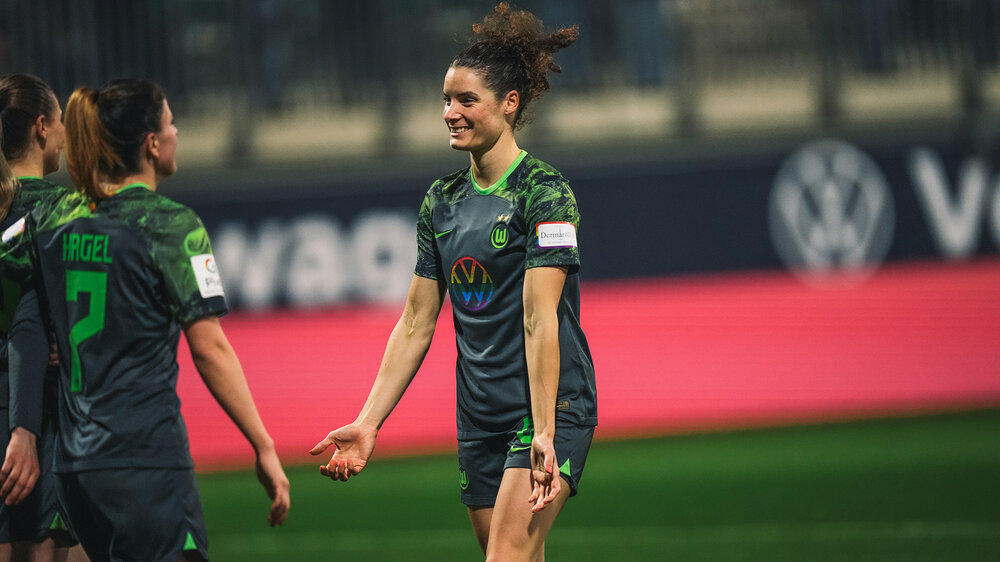 VfL-Wolfsburg-Spielerin Dominique Janssen streckt die Arme in Richtung ihrer Mitspielerinnen aus und lacht dabei.
