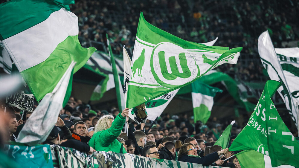 Fans vom VfL Wolfsburg schwenken im Fanblock die Vereinsfahnen.