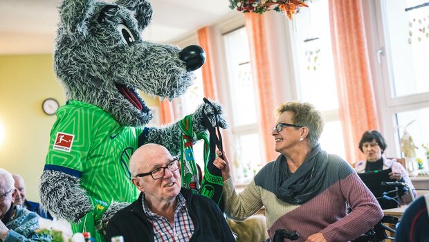 VfL-Wolfsburg-Maskottchen Wölfi übergibt einer AWO-Altenheim-Bewohnerin eine Tasche.
