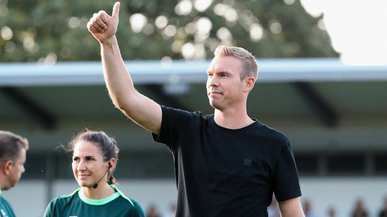 Trainer der VfL-Wolfsburg-Frauen Tommy Stroot steht auf dem Fußballplatz. Er streckt seinen Arm in die Luft und zeigt zeigt mit dem Daumen hoch.