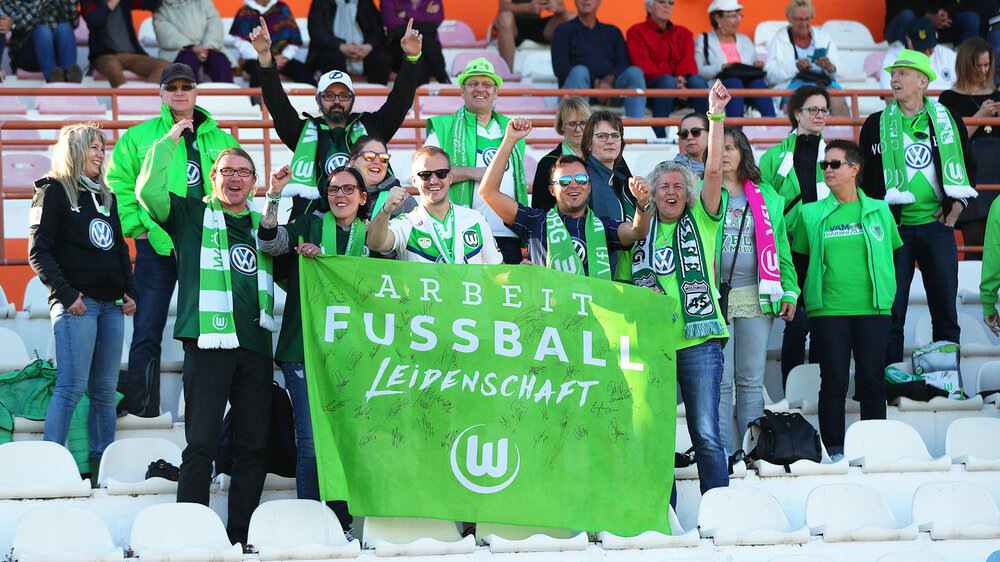 VfL Wolfsburg-Fans posieren in Fankleidung mit Fahne auf den Rängen des AOK Stadions.