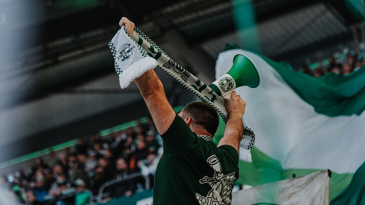 Ein Fan des VfL Wolfsburg hält einen grün-weißen Schal und ein Mikrofon hoch.
