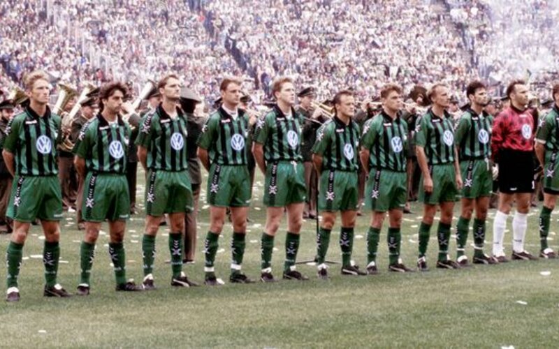 Die Herrenmannschaft des VfL Wolfsburg von 1994 steht auf dem Spielfeld mit einer Parade im Hintergrund.