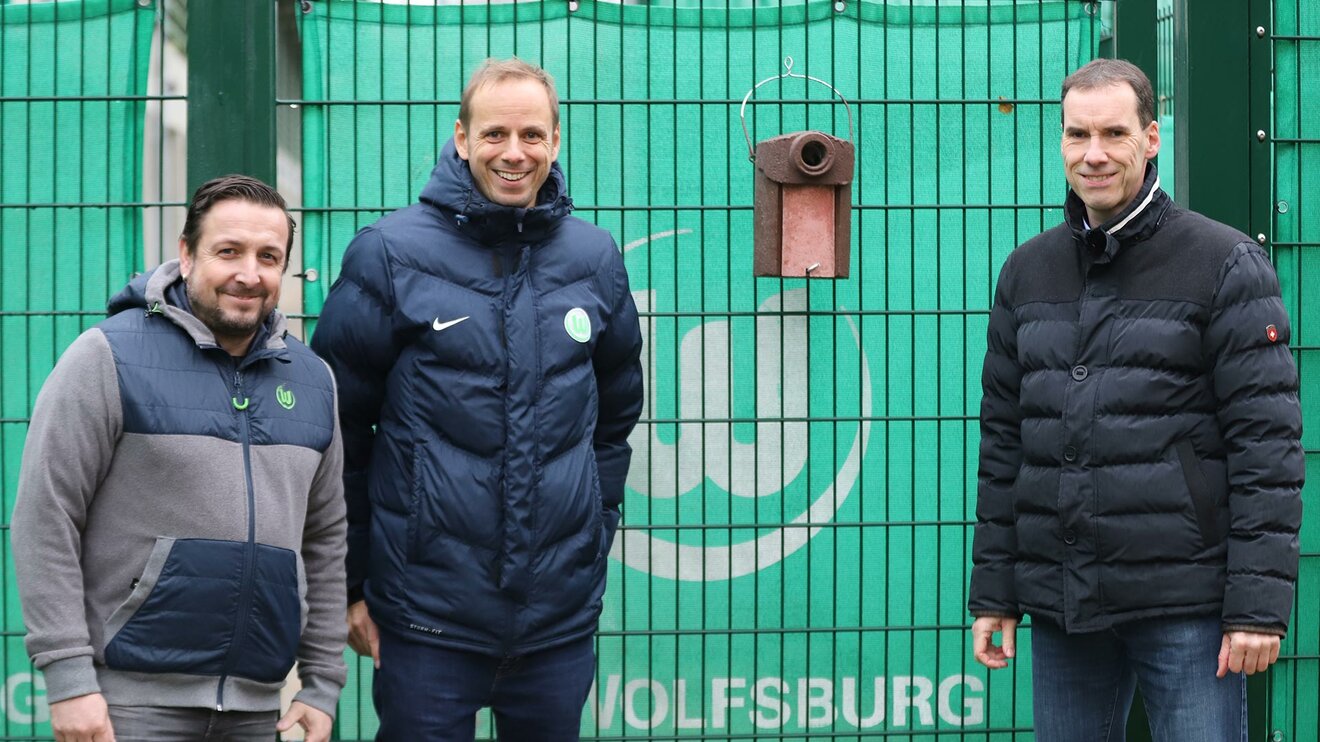 Zwei VfL- und ein NABU-Vertreter stehen vor einem Zaun zu einem Trainingsgelände des VfL-Wolfsburg, an dem ein Nistkasten für Stare angebracht ist.