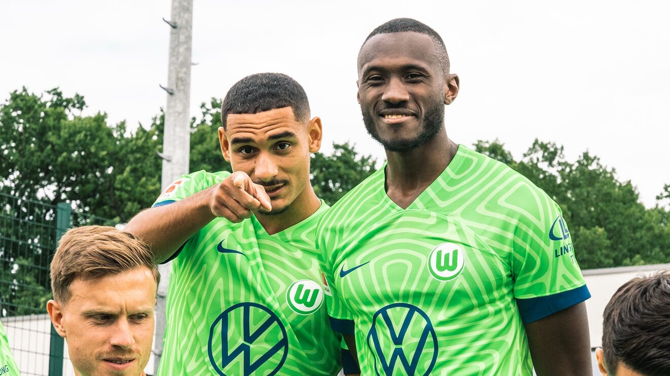 VfL-Wolfsburg-Spieler Maxence Lacroix steht neben seinem ehemaligen Teamkollegen Josh Guilavogui und zeigt mit dem Finger auf die Kamera.