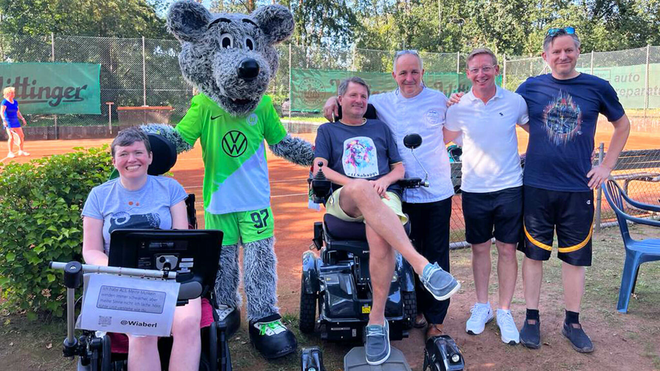 VfL Wolfsburg Maskottchen Wölfi posiert im Rahmes eines Charity Events mit VfL Funktionären und Fans im Rollstuhl.