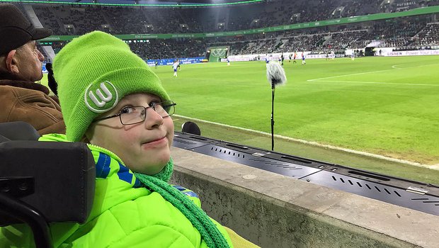 Ein junger Fan vom VfL Wolfsburg sitzt nah am Spielfeld und schaut in die Kamera.