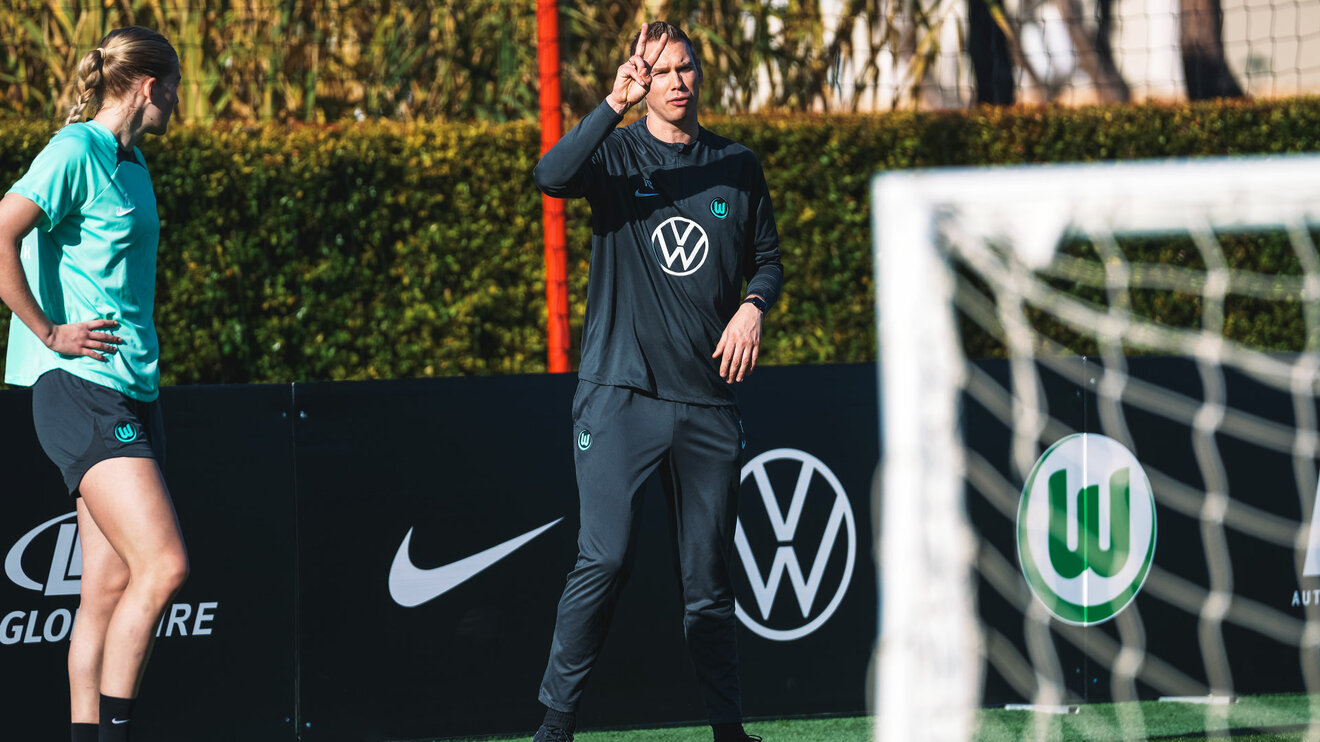Der Trainer der VfL Wolfsburg Frauen, Tommy Stroot, steht auf dem sonnigen Trainingsplatz in Portugal und zeigt den Spielerinnen eine zwei mit den Fingern.