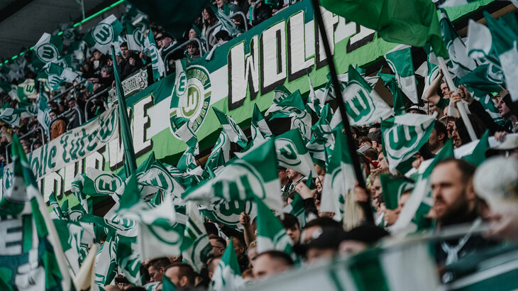Fans des VfL Wolfsburg schwenken grün-weiße Fahnen auf der Tribüne.