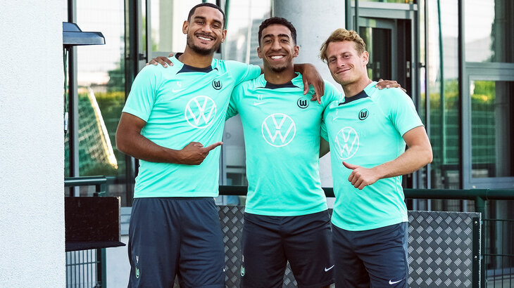 VfL Wolfsburg Spieler Lacroix, Tomas und Cozza posieren Arm in Arm im Trainingsdress.