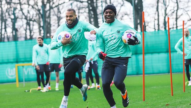 Lukas Nmecha und Ridle Baku vom VfL Wolfsburg laufen jeweils mit einem Ball in der Hand den Parcours und lachen.
