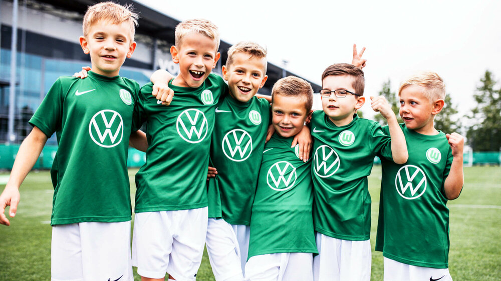 Kleine Fußballfans im VfL Trikot posieren Arm in Arm auf dem Trainingsgelände vor der Volkswagen Arena.