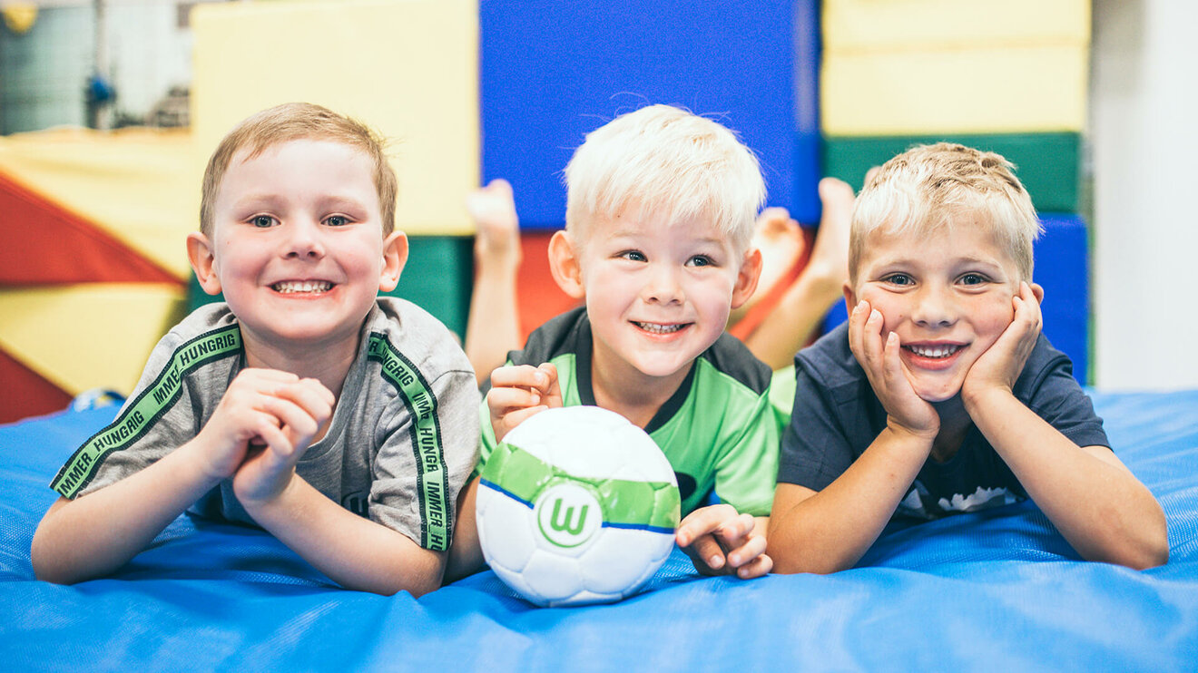 Drei Kinder liegen auf einer Matte, davor ein Ball des VfL Wolfsburg.