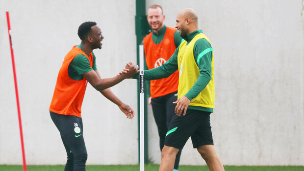 Lukebakio und Brooks schlagen lachend miteinander ein, im Hintergrund steht Arnold und schaut den beiden grinsend zu beim Training des VfL Wolfsburg an der Volkswagen Arena.