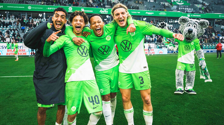 Die VfL Wolfsburg Spieler Tomas, Paredes Vranckx und Bornauw stehen nebeneinander Arm in Arm auf dem Spielfeld und jubeln in die Kamera.
