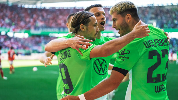 Die VfL Wolfsburg-Spieler Lovro Majer, Yannick Gerhardt und Jonas Wind stehen Arm in Arm auf dem Platz und freuen sich.