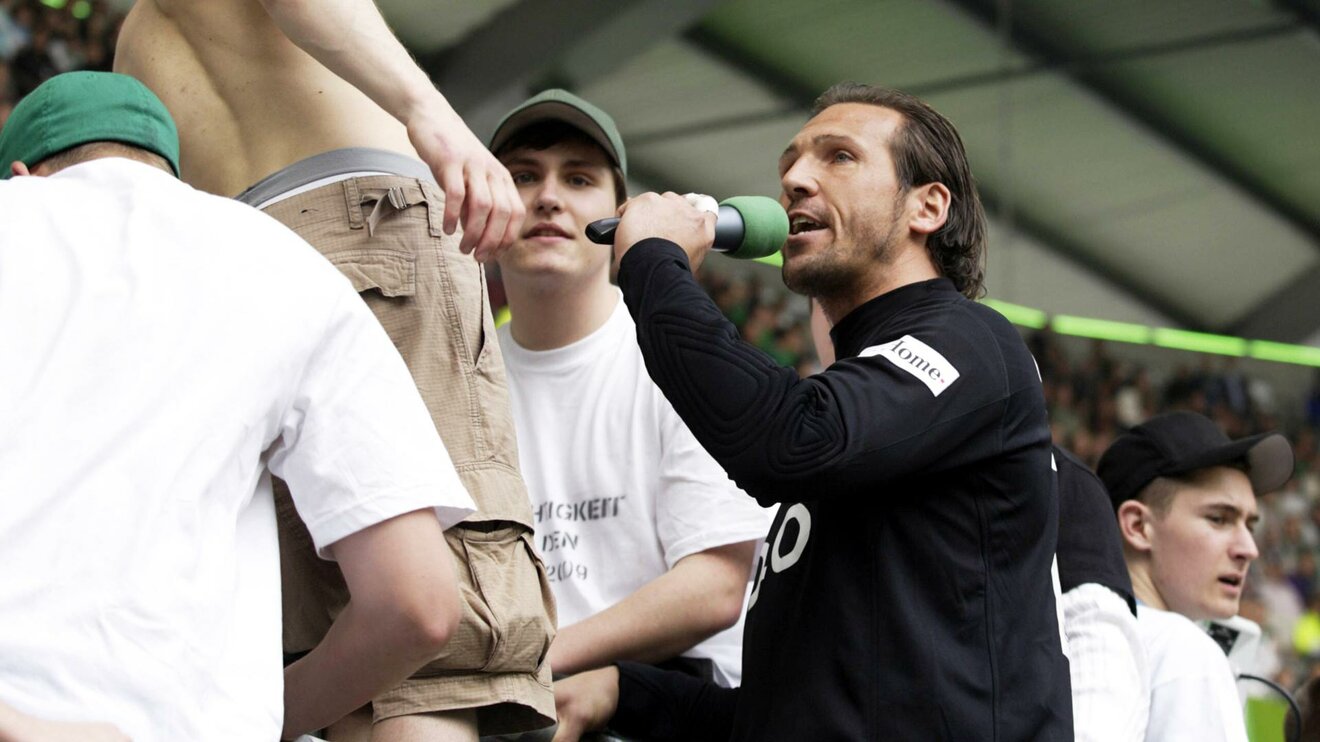 Der ehemalige Torwart des VfL Wolfsburg Andre Lenz steht mit einem Mikrofon in der Hand in der Nordkurve.