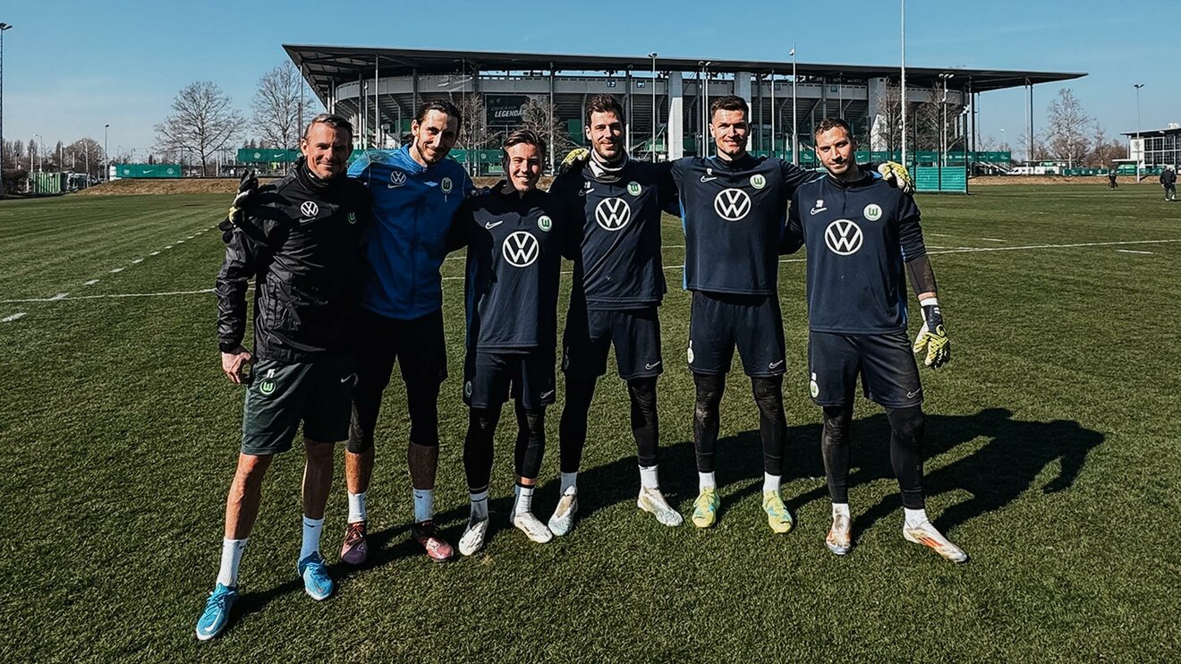 Die VfL-Wolfsburg-Torhüter Kamil Grabara, Marius Müller, Pavao Pervan und Niklas Klinger stehen Arm in Arm mit Torwart-Influencer Michael Bolvin und Torwarttrainer Pascal Formann auf dem Trainingsplatz vor der Volkswagen Arena.