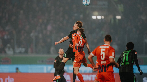VfL-Wolfsburg-Spieler Kevin Behrens und ein Gegenspieler springen für einen Kopfball in die Höhe.