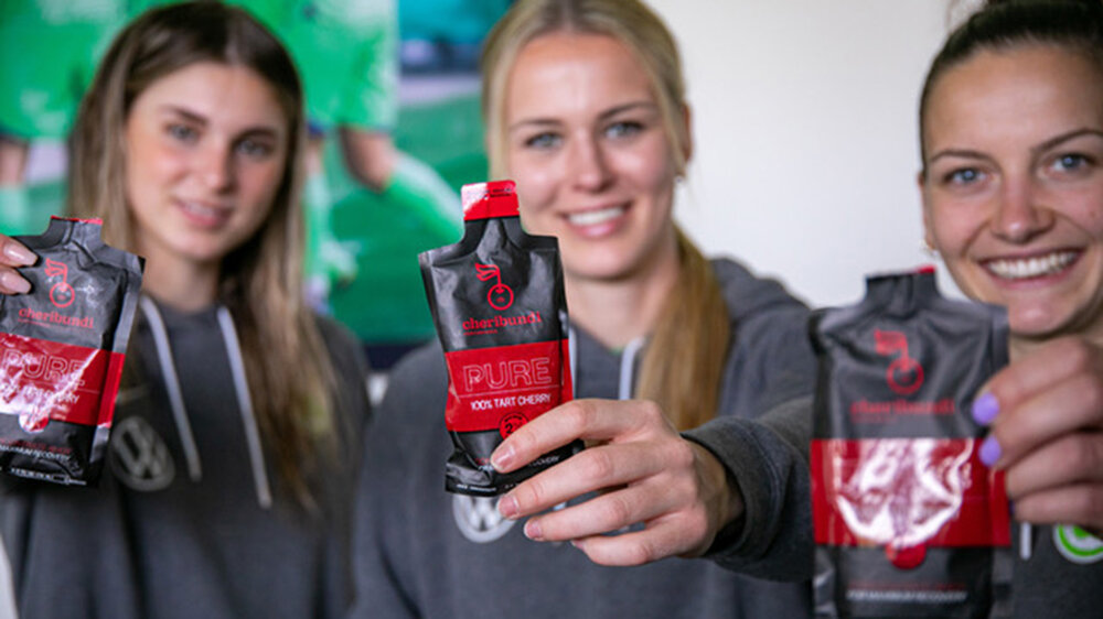 Die Spielerinnen des VfL Wolfsburg Jule Brand, Merle Frohms und Joelle Wedemeyer präsentieren die Produkte von Cheribundi in die Kamera.