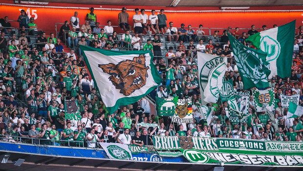 Die Fans des VfL Wolfsburg stehen auf der Tribüne und schwenken Fahnen.