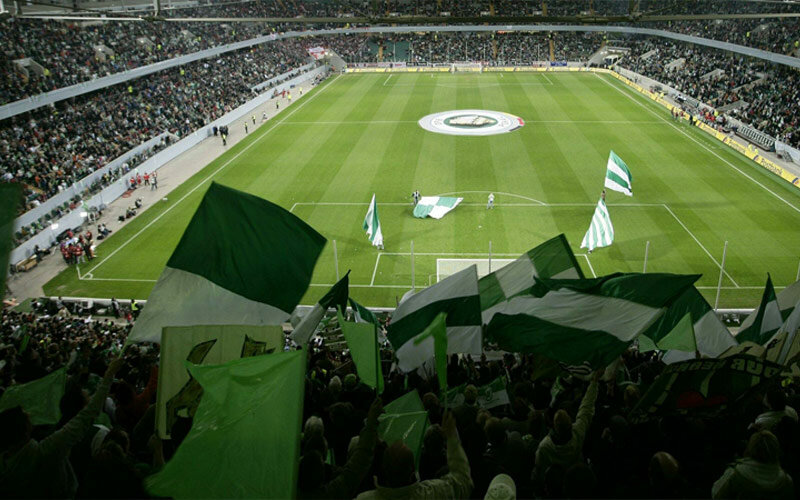 Ein Foto von der Volkswagen Arena vom VfL Wolfsburg mit jubelnden Fans mit Fahnen in der Nordkurve im Vordergrund.