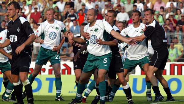 Mehrere Spieler vom VfL-Wolfsburg und von Gladbach stehen aufgereiht und blicken gespannt nach dem Ball. Ein VfL-Wolfsburg-Spieler wird dabei von einem Gladbach-Spieler, der hinter ihm steht, von hinten umarmt und festgehalten.