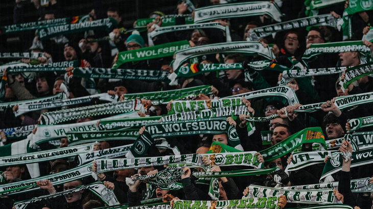 Fans des VfL Wolfsburg halten ihre Schals hoch und stehen auf der Tribüne.