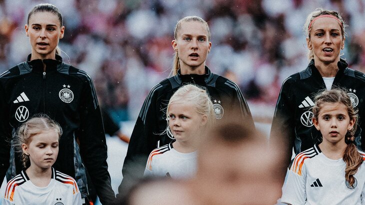 Die drei VfL-Wolfsburg-Spielerinnen Jule Brand, Janina Minge und Kathy Hendrich steht in Trainingsjacken der Deutschen Nationalmannschaft nebeneinander und vor ihnen stehen Einlaufkinder.