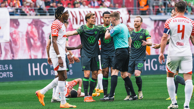 Ein VfL-Wolfsburg-Spieler steht vor dem Schiri und hebt seine Arme links und rechts ausgestreckt auf Schulterhöhe. Neben ihm stehen Mitspieler und  Spieler von RB Leipzig. Ein RB Leipzig Spieler liegt hinter ihm auf dem Boden. 