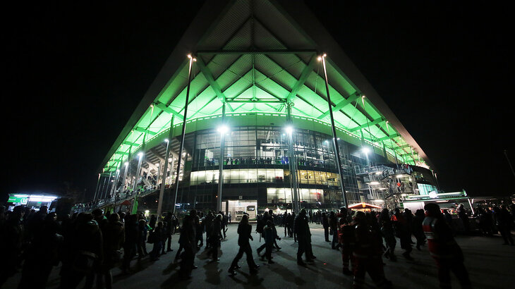 Fans des VfL Wolfsburg laufen in der Dunkelheit vor der grün beleuchteten Volkswagen Arena.