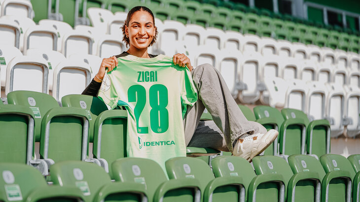 Cora Zicai sitz auf der Tribüne und hält ein VfL-Wolfsburg-Trikot mit der Rückennummer 28 hoch.