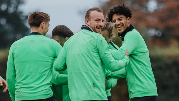U19-Kapitän Jonathan Akaegbobi und Maximilian Arnold vom VfL-Wolfsburg, tragen beide grünen Trainingsjacken und stehen im Kreis mit anderen Spielern. Die beiden drehen sich in Richtung Kamera um und lächeln.  