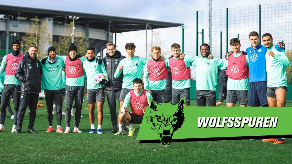 Die Lizenzmannschaft des VfL Wolfsburg auf dem Trainingsplatz, davor befindet sich eine Grafik mit der Aufschrift "Wolfsspuren".